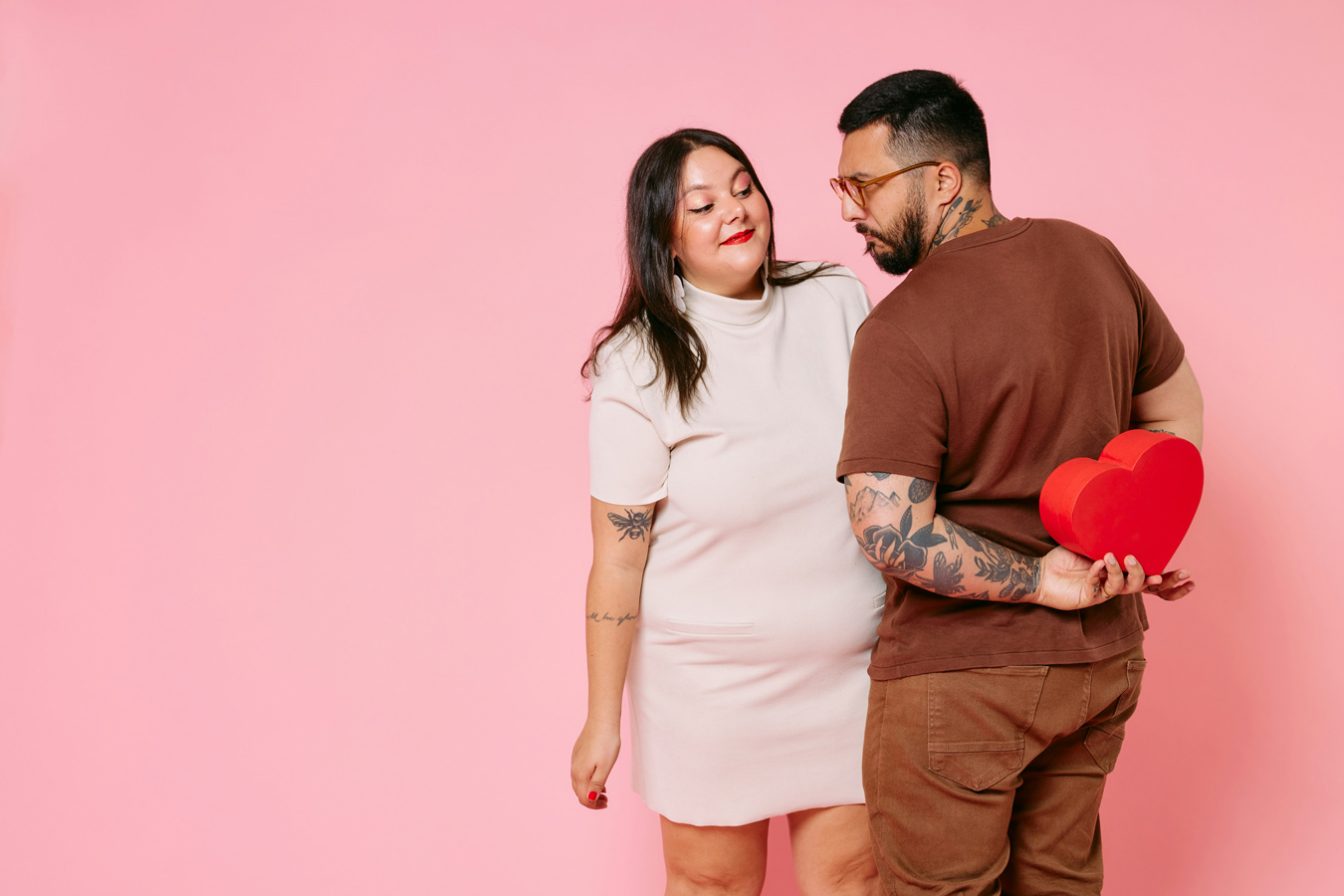 A woman in a cream dress looks lovingly at a man with glasses and tattoos, who is hiding a red heart-shaped box behind his back against a pink background. The man is looking back at the woman with a slight smile.