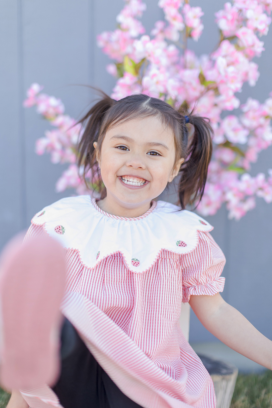 A young girl with pigtails smiles brightly, wearing a red and white striped dress with a white collar embroidered with strawberries. Behind her is a flowering tree with pink blossoms.