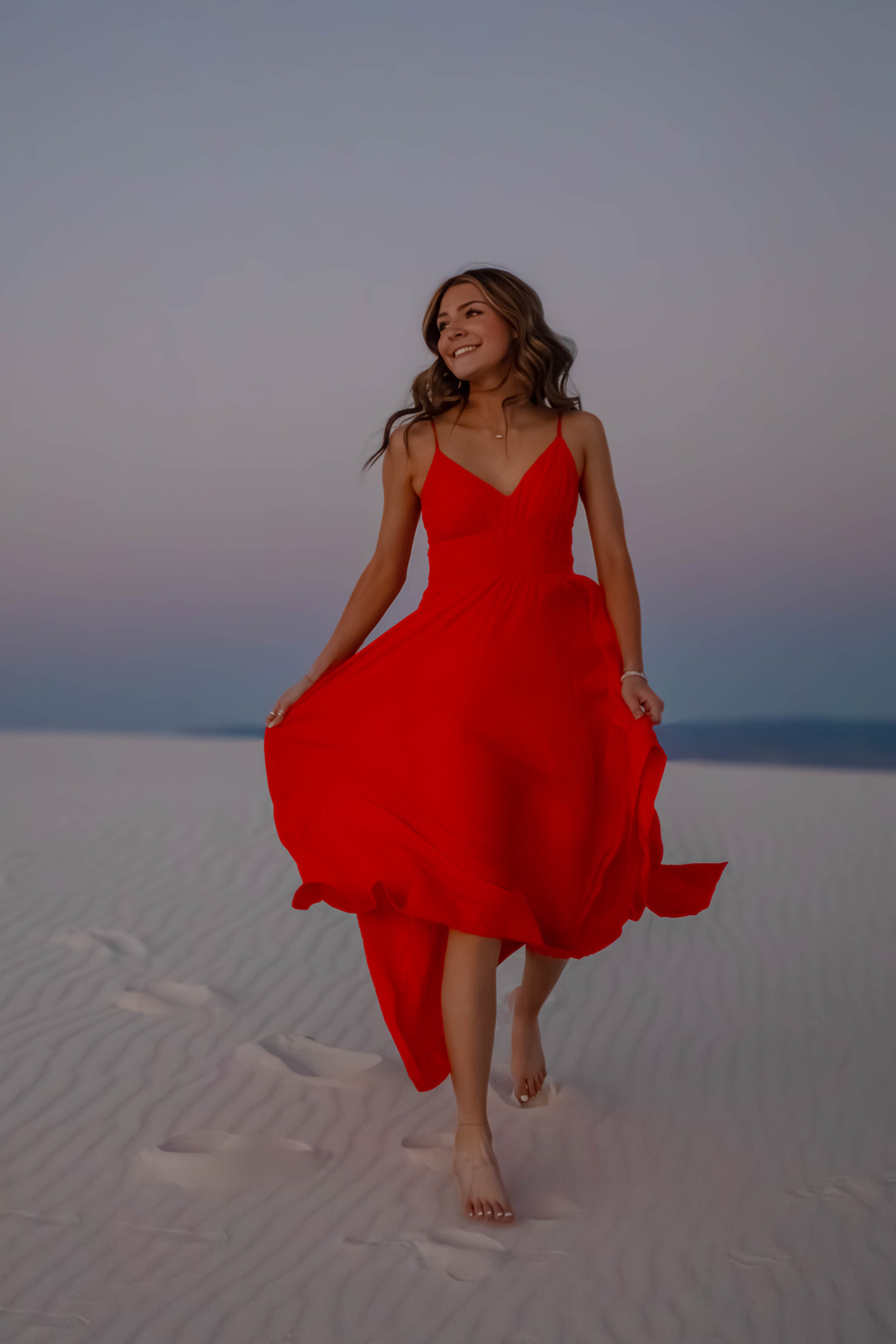 A young woman with long brown hair smiles and walks barefoot across white sand, wearing a flowing red dress with spaghetti straps. The sky is a soft gradient of blue and pink in the background. Photo by Cassidee Wiemers