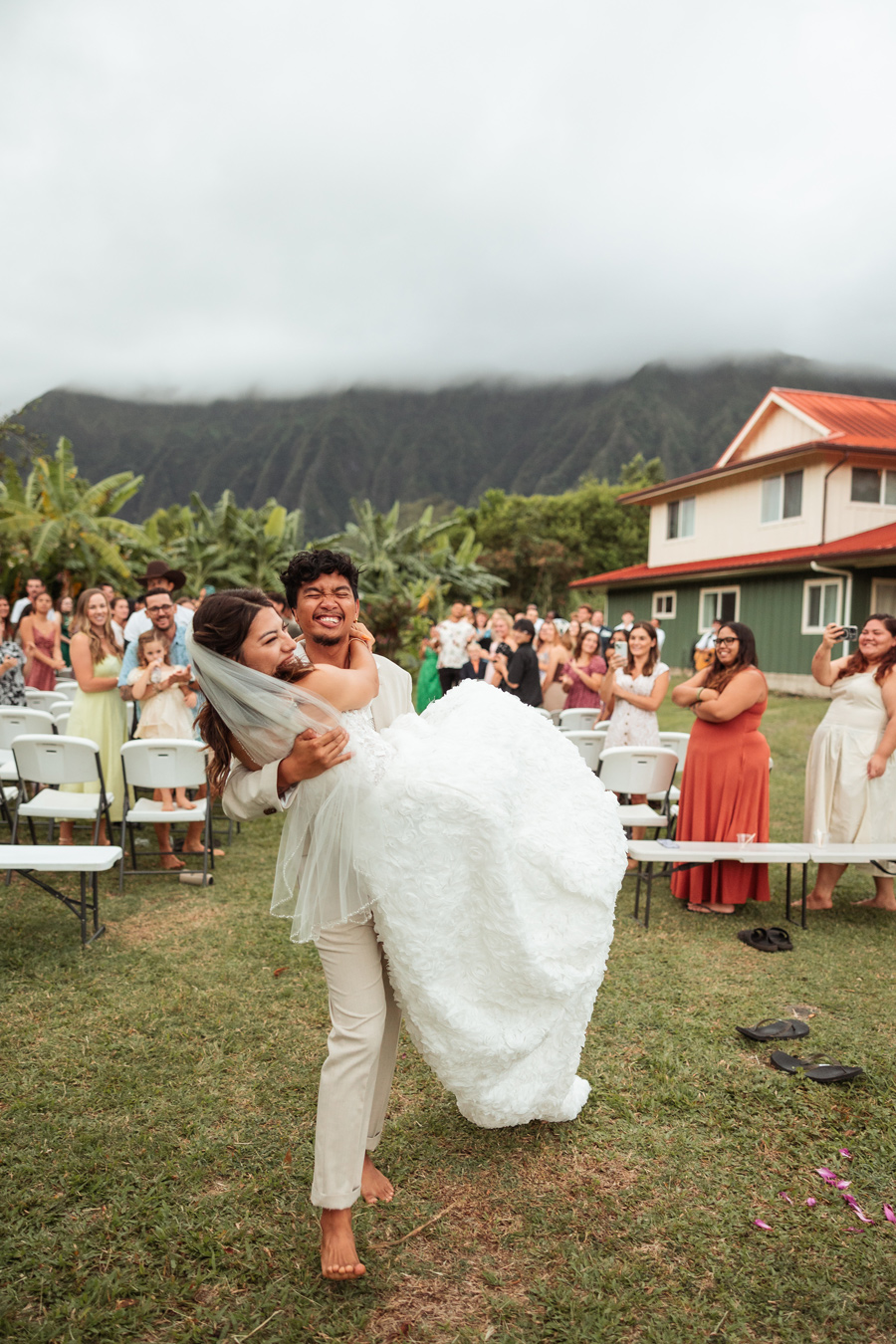 A groom in a tan suit and barefoot carries his bride in a white gown in front of wedding guests, with a foggy mountain range in the background. The bride and groom are smiling and laughing. Photo by Daphne Scott Photography.