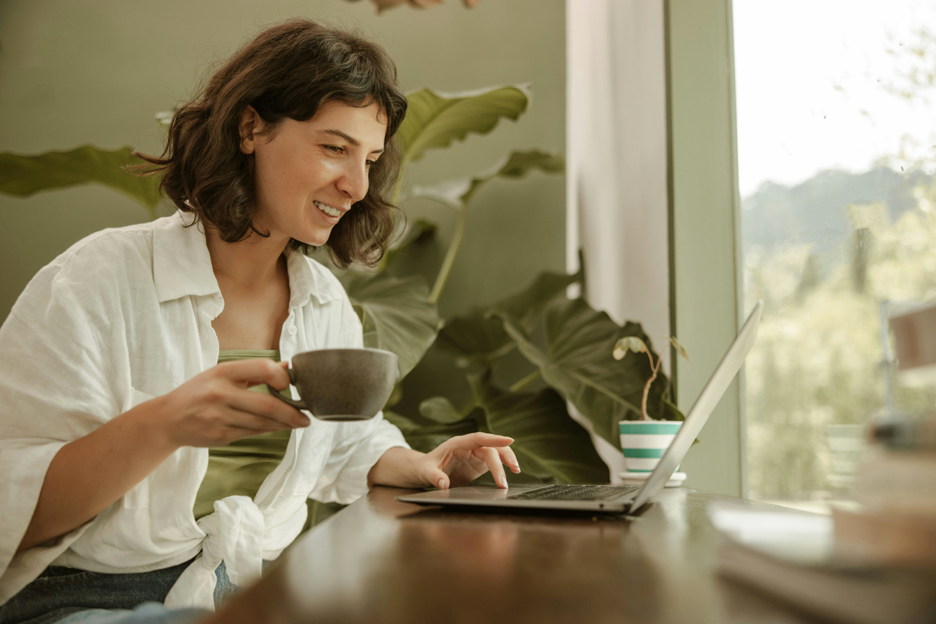 A smiling woman with short brown hair sits at a wooden table, holding a mug and using a laptop, with large green plants and a window in the background. She is wearing a white shirt tied at the waist.
