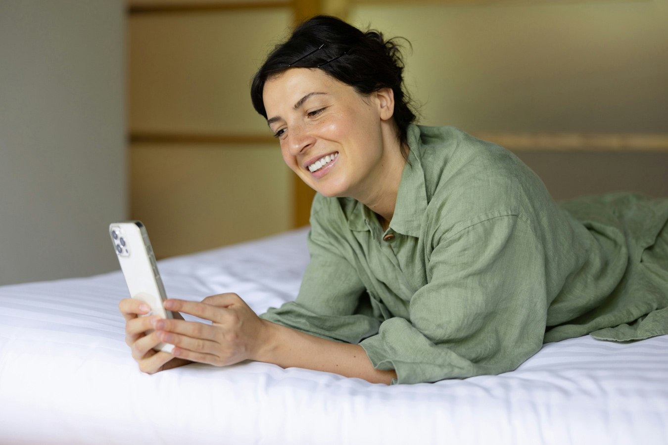 A smiling woman in a green linen shirt and pants lies on a white bed, looking at her smartphone. Her dark hair is pulled back with bobby pins.