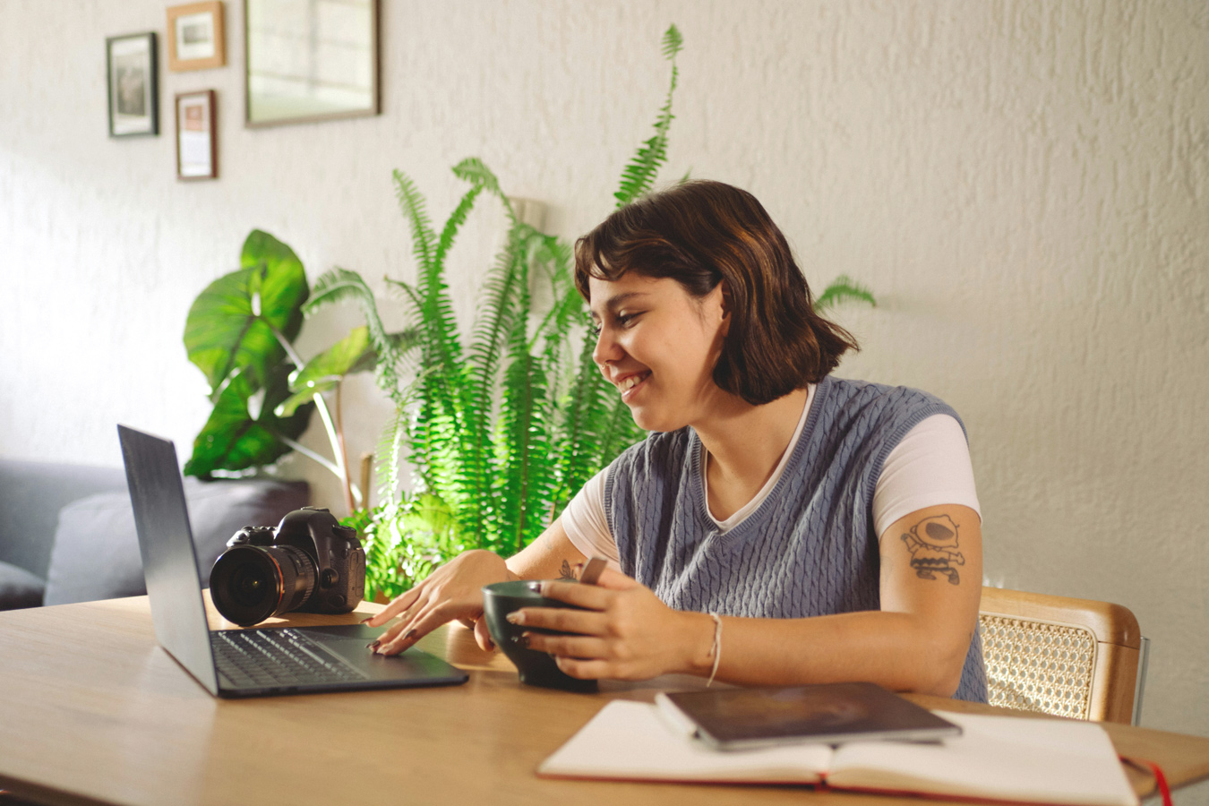 A smiling woman with short brown hair and a tattoo on her arm sits at a wooden table, using a laptop with a camera next to it, and holding a mug. Behind her are several plants and framed pictures on the wall.