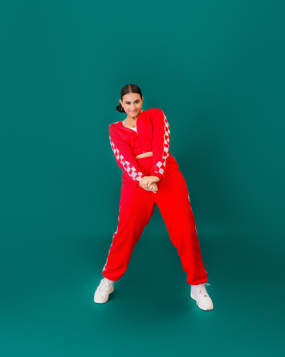A young dancer with dark hair pulled back into a bun poses in a red tracksuit with white checkered stripes down the arms and legs for a hip hop routine, paired with white sneakers, against a teal background. She is smiling slightly with her hands clasped in front of her. Photo by JSPS Schools.