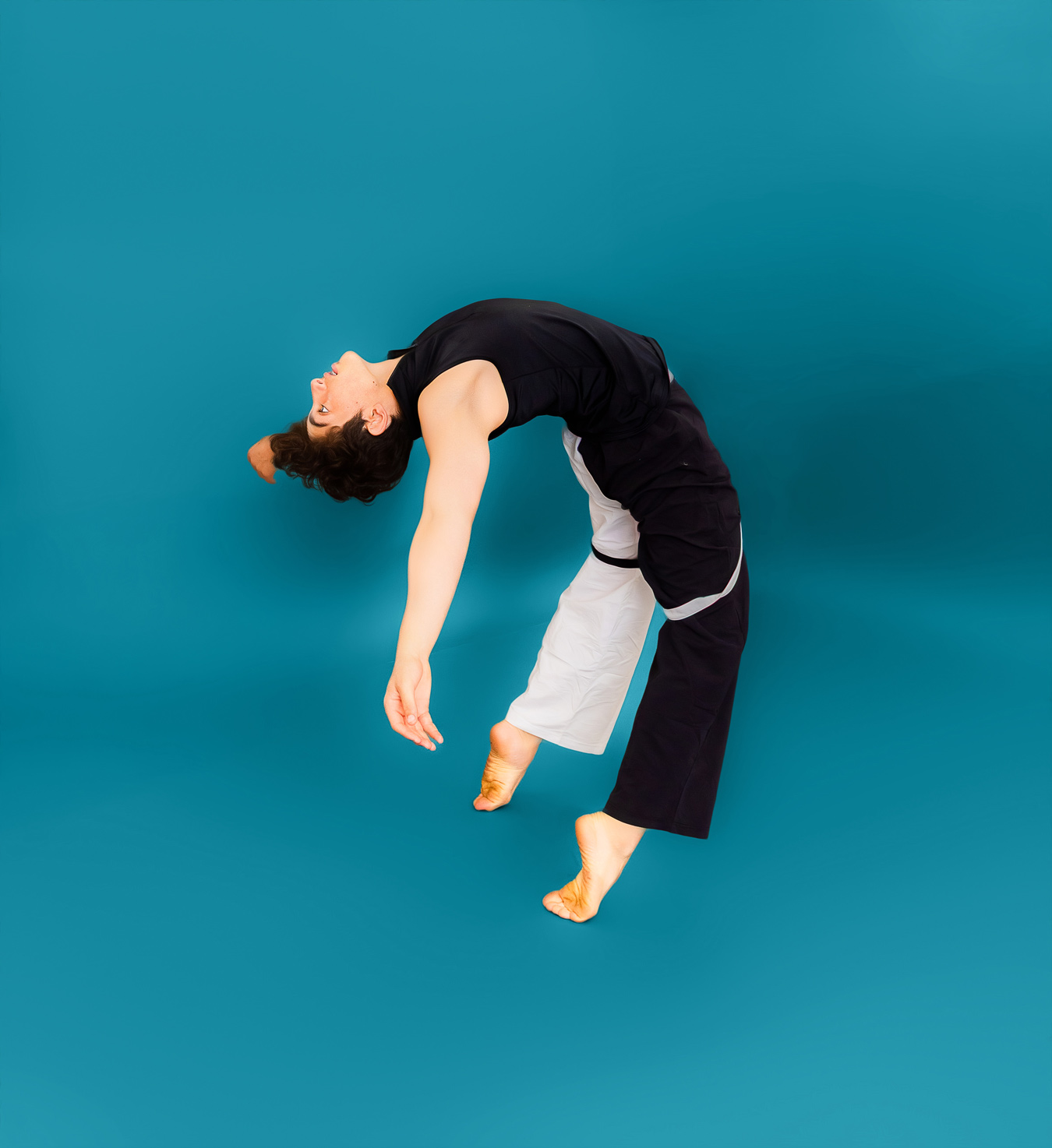 A dancer in a black top and black and white pants arches backward against a teal background. The dancer's head is tilted back, and their arms and feet are extended. Photo by JSPS Schools.