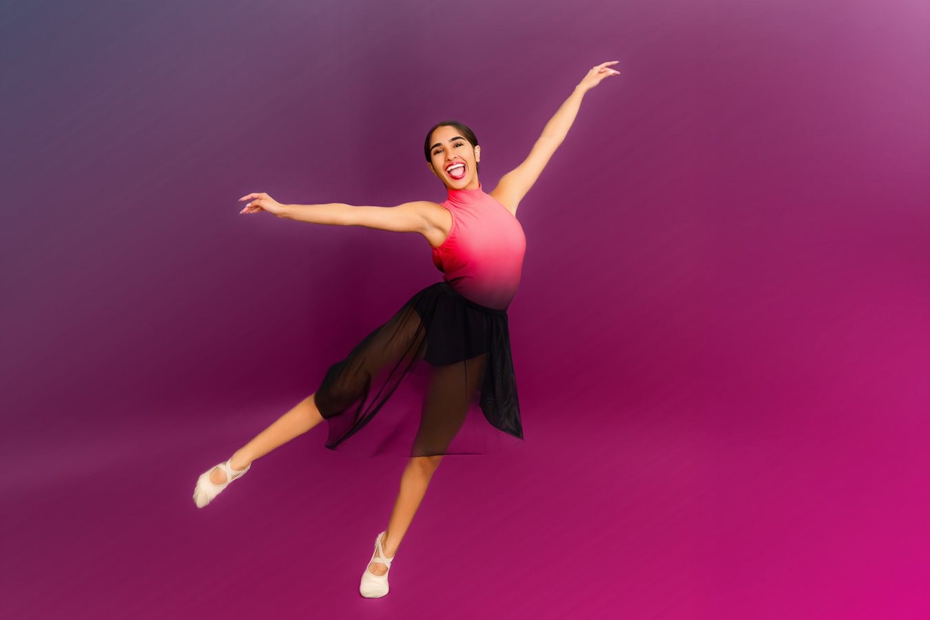 A smiling dancer in a pink and black leotard and black skirt leaps against a gradient purple background, arms outstretched and legs extended. She wears white ballet shoes. Photo by JSPS Schools.