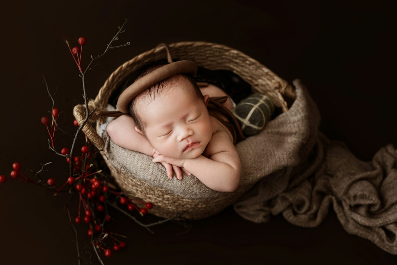 A newborn baby wearing a small brown hat sleeps peacefully in a woven basket lined with a soft, neutral-toned blanket. A sprig of red berries adorns the side of the basket, set against a dark brown background.