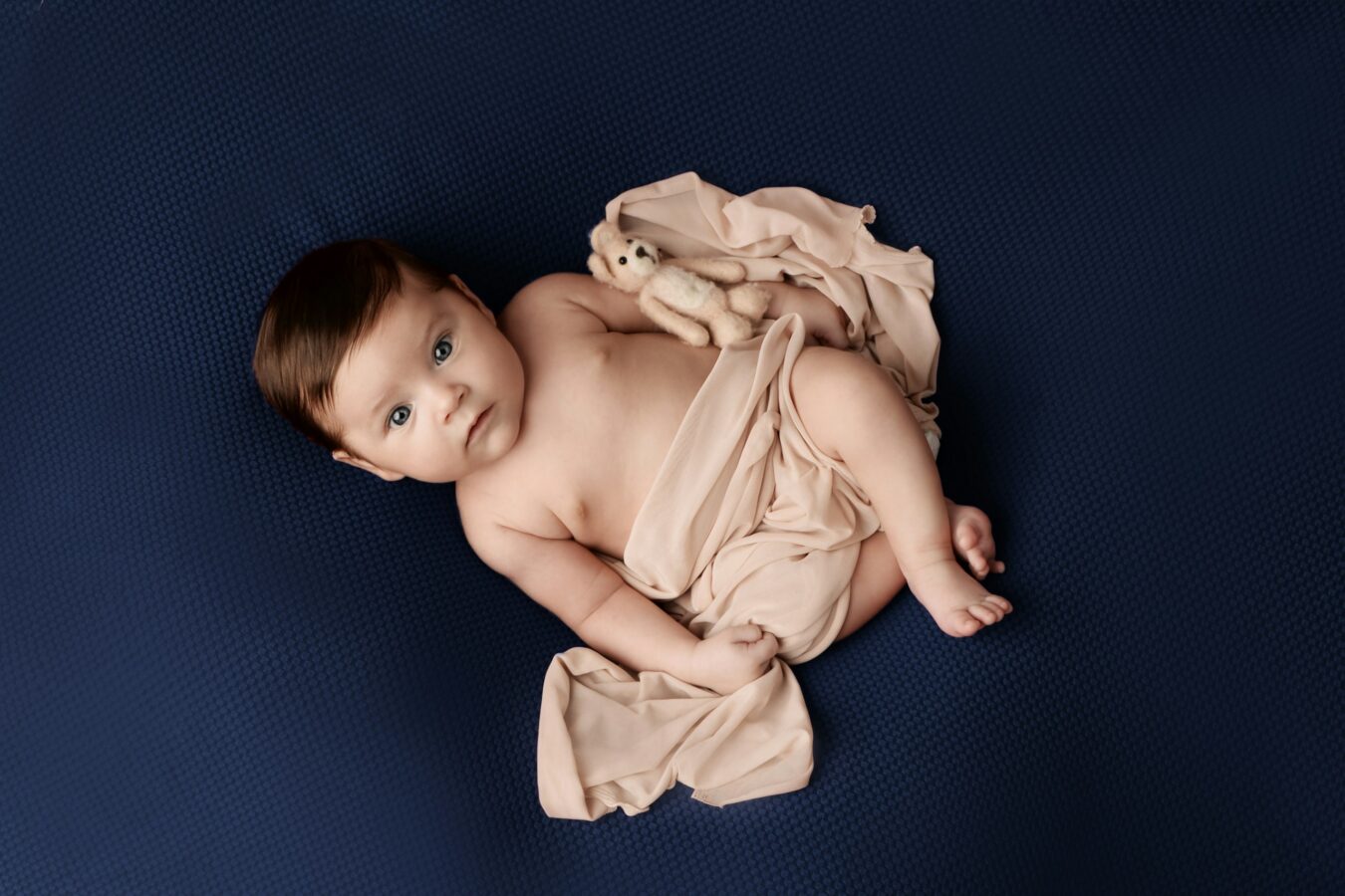 A baby with brown hair and blue eyes lies on a dark blue textured blanket, partially wrapped in a light peach-colored fabric, with a small teddy bear resting on their side. The baby is looking directly at the viewer with a curious expression. Lighting was provided by a strobe or flash.