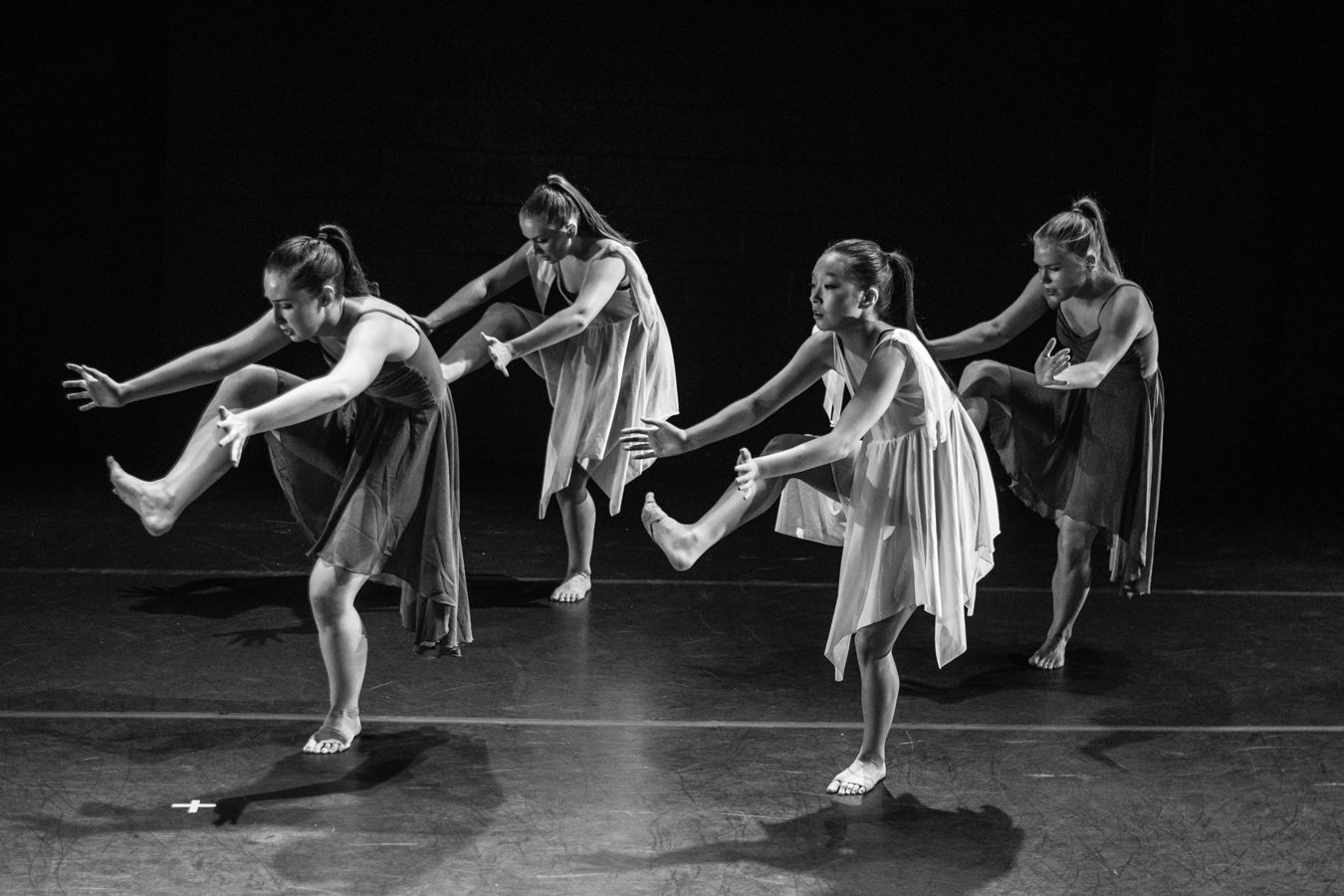 Four barefoot dancers in flowing dresses are captured mid-performance in a black and white photograph, each with one leg extended and arms outstretched in a synchronized pose. The dancers are positioned on a dark stage with minimal lighting, creating dramatic shadows.