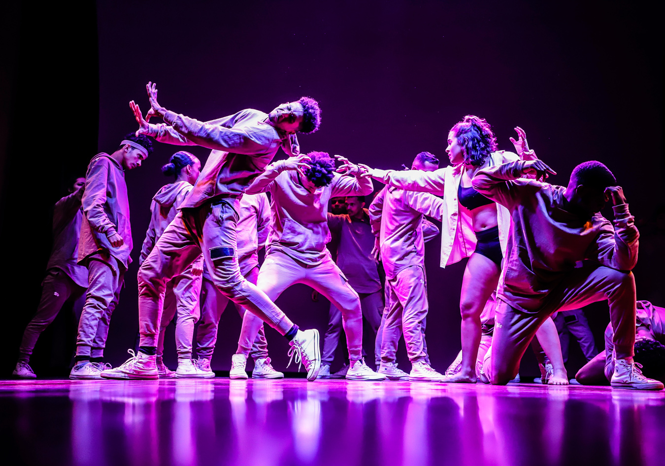 A group of dancers in casual clothing perform on a stage lit with vibrant purple light, their movements dynamic and synchronized. One dancer stands out in a black bra and shorts with an open white shirt, adding a striking contrast to the otherwise uniformly dressed group.