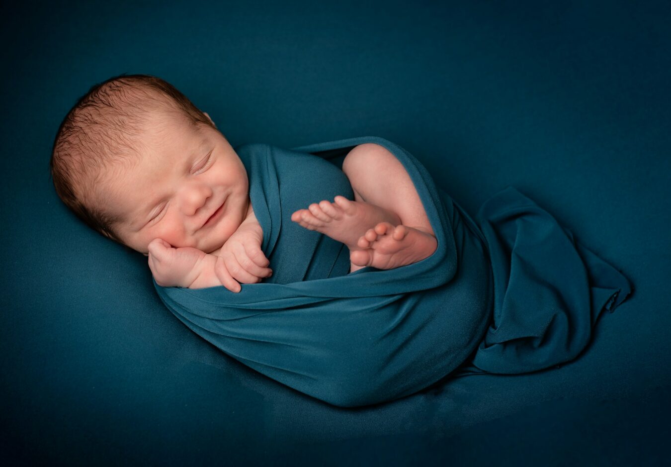A newborn baby with closed eyes and a slight smile is swaddled in a teal fabric against a matching teal background. The baby's tiny hands and feet are visible, adding to the image's tenderness. Photo by Adele Morris Photography via Unsplash.