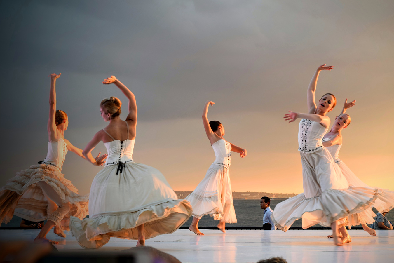 Five ballerinas in white dresses and corsets gracefully dance on an outdoor stage against a backdrop of a sunset sky and distant coastline. Their arms are raised in unison, creating a dynamic and elegant composition.