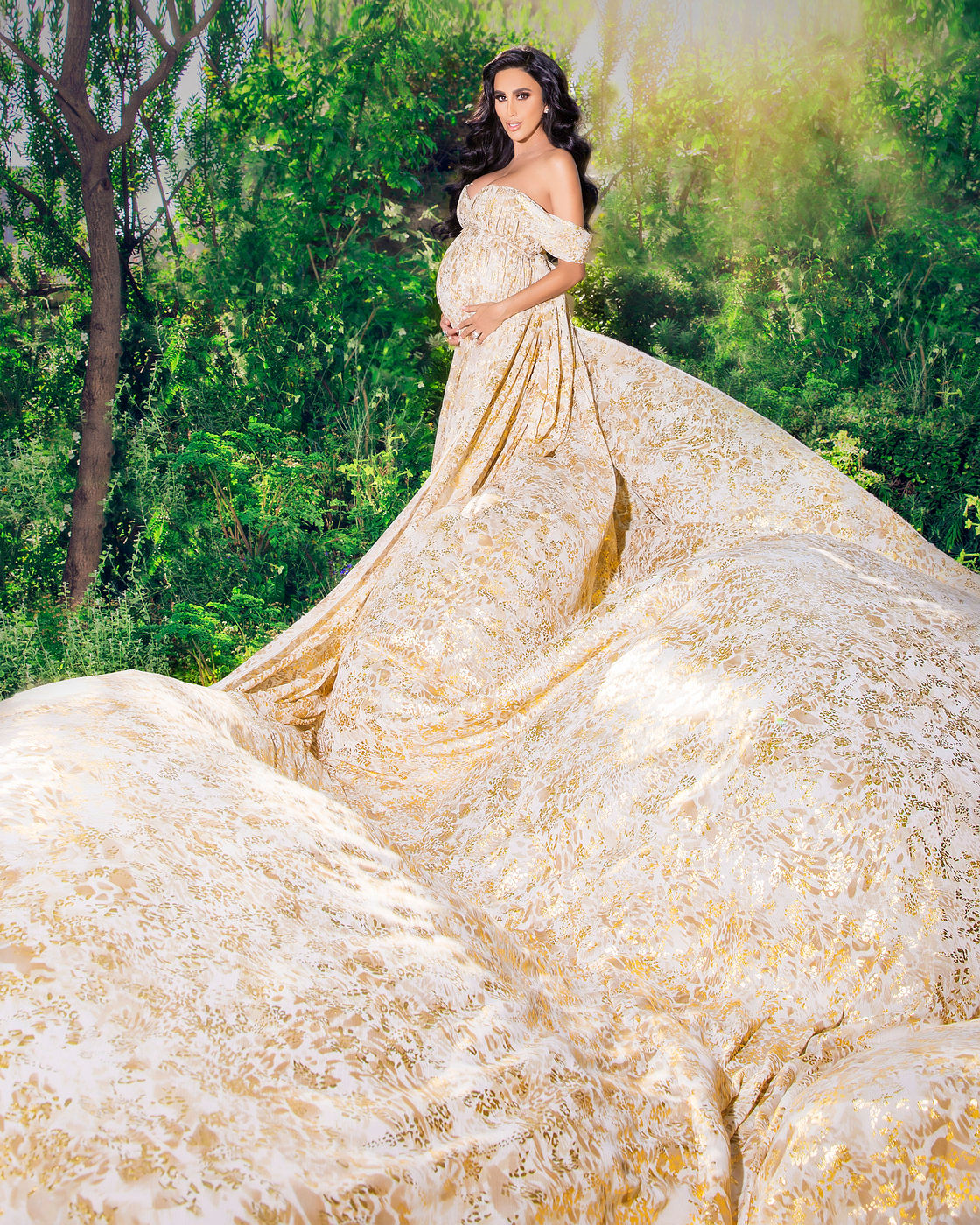A pregnant woman with long dark hair stands in a forest, wearing an off-the-shoulder gown with a long, flowing train of white and gold fabric. She is holding her baby bump with both hands. Photo by Adam Opris.
