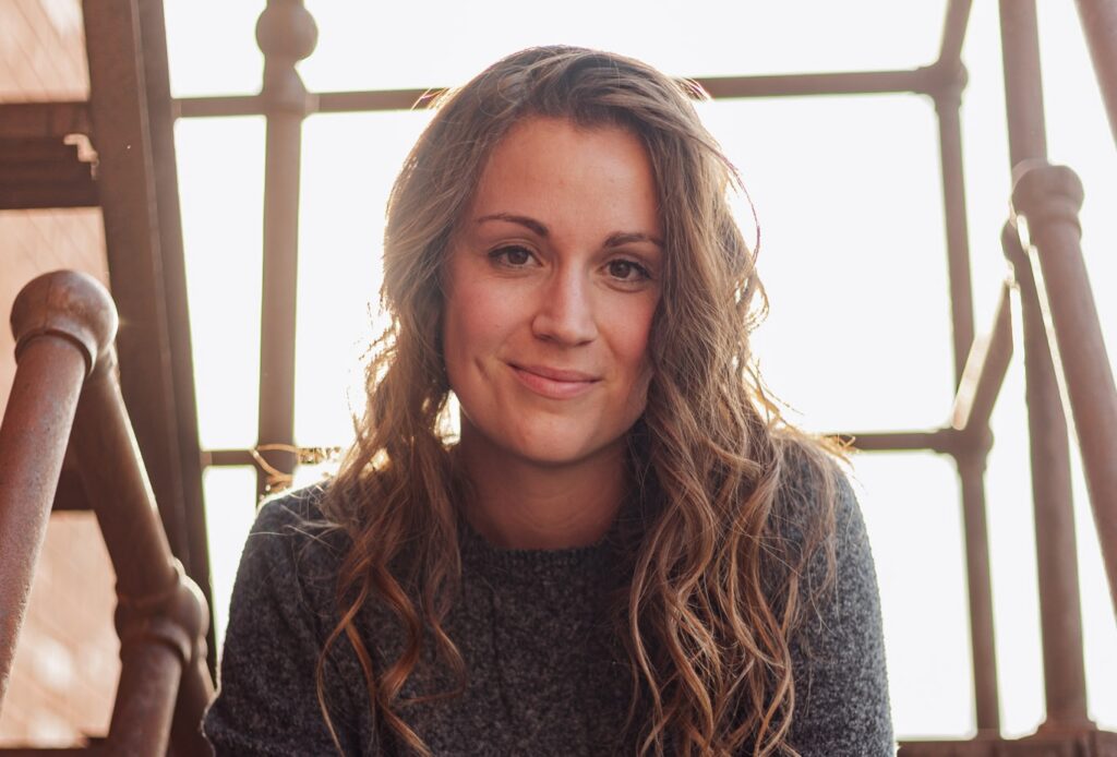 A smiling woman with long, wavy brown hair sits in front of a rusty metal staircase. She wears a gray sweater. Sunlight shines through the stairs behind her.