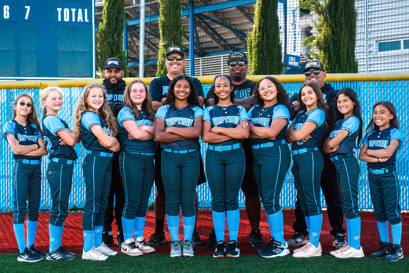 A softball team, Riptide, poses with coaches in front of a blue fence and scoreboard reading 6 7 TOTAL. The team wears matching blue and black uniforms with the team name on the front. Coaches wear Riptide apparel.