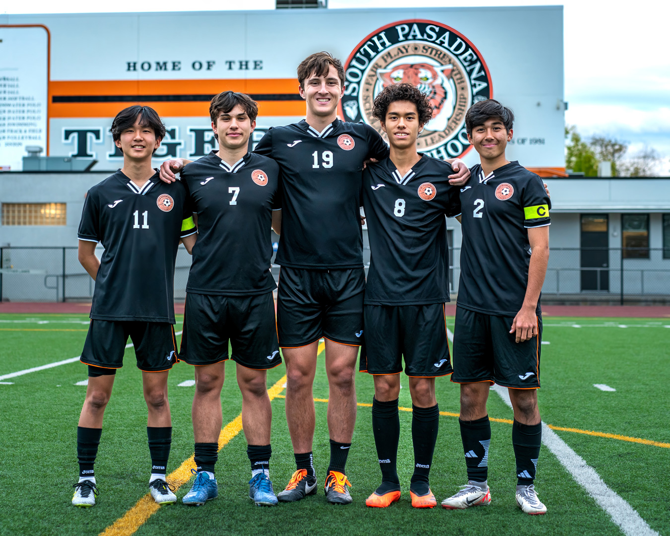 Five soccer players in black uniforms with orange trim stand on a green field. The player on the right wears a captain's armband. Behind them is a building with the words HOME OF THE SOUTH PASADENA TIGERS.