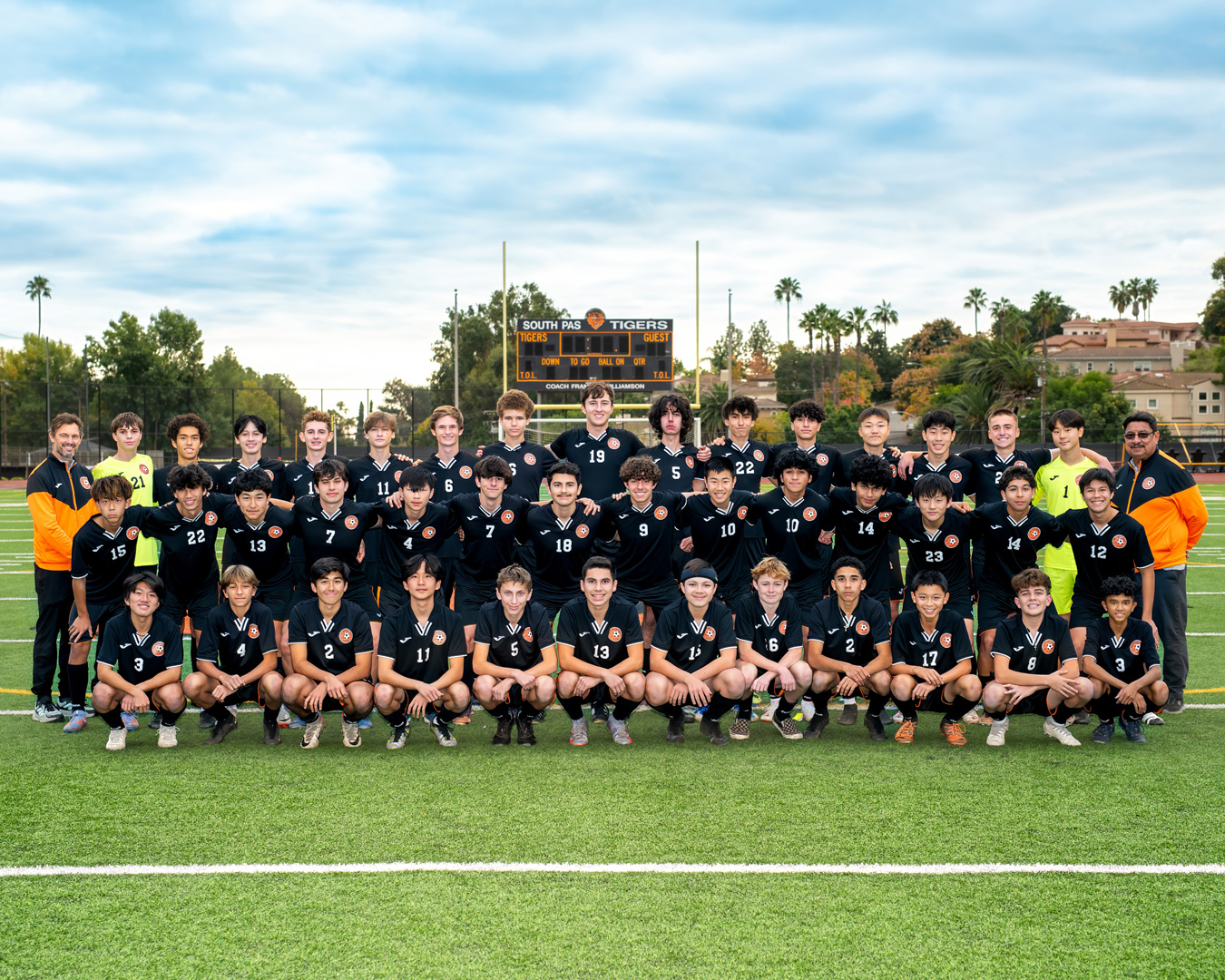 A soccer team poses for a photo on a green field in front of a scoreboard that reads SOUTH PAS TIGERS. The team wears black jerseys with orange accents and numbered white text. Two coaches stand on either side of the team.