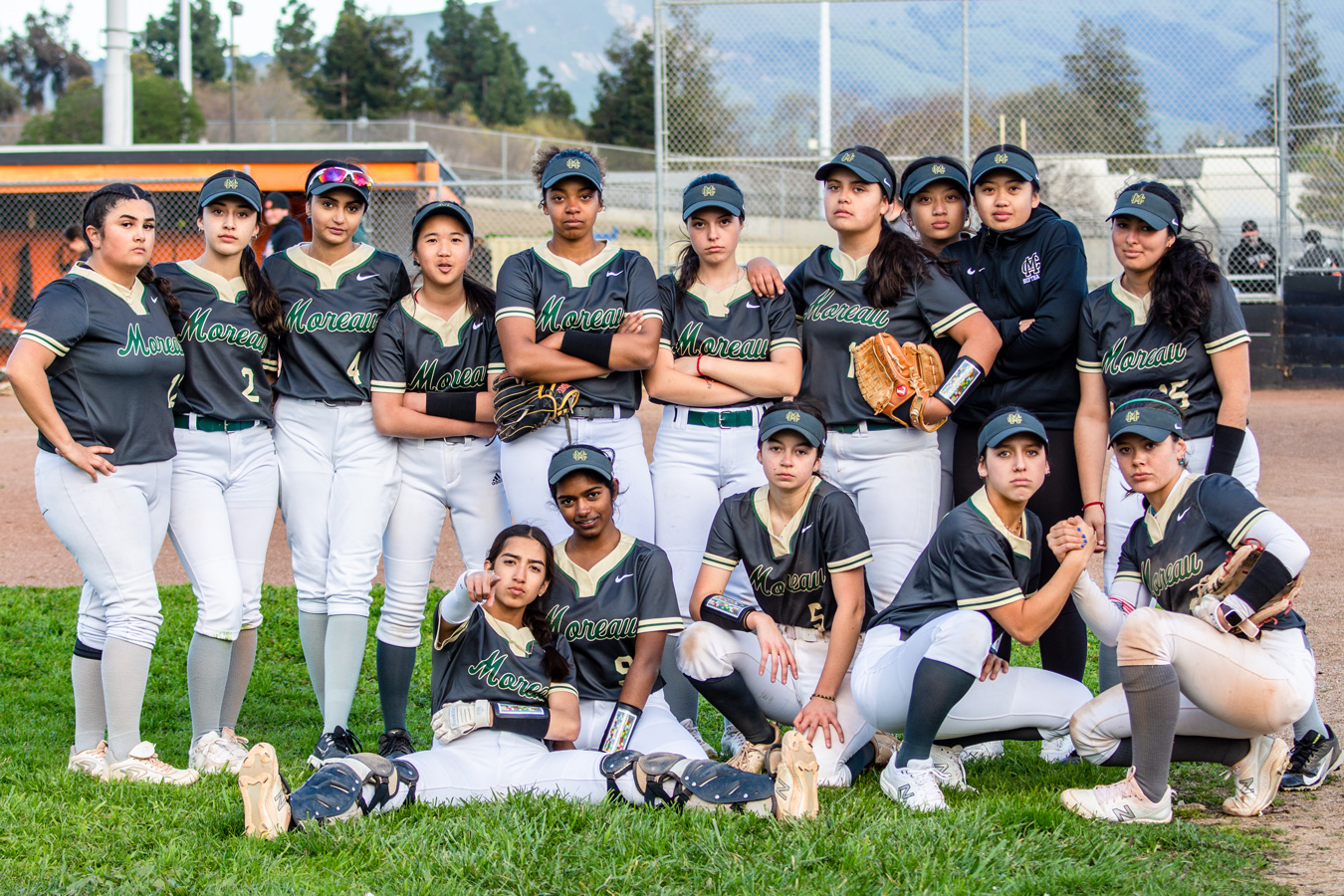 A softball team, wearing gray and white uniforms with Moreau printed on the front, poses for a team photo on a field. Mountains are visible in the background.