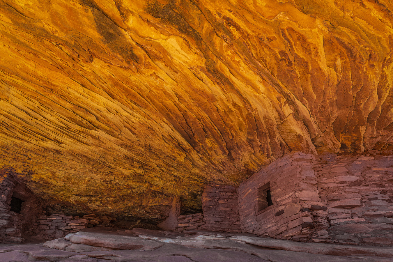 Ancient stone ruins of the House on Fire cliff dwelling sit nestled beneath a massive, swirling orange and yellow sandstone overhang that resembles flickering flames.
