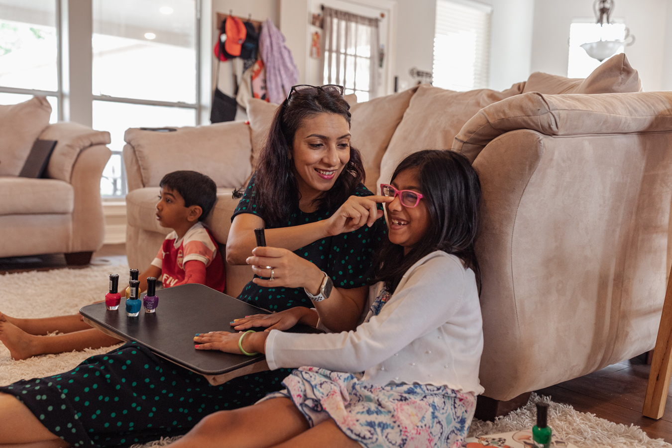 A woman in a green polka-dot dress sits on a living room floor, playfully touching the nose of a young girl in pink glasses who is getting her nails painted, while a young boy sits in the background.