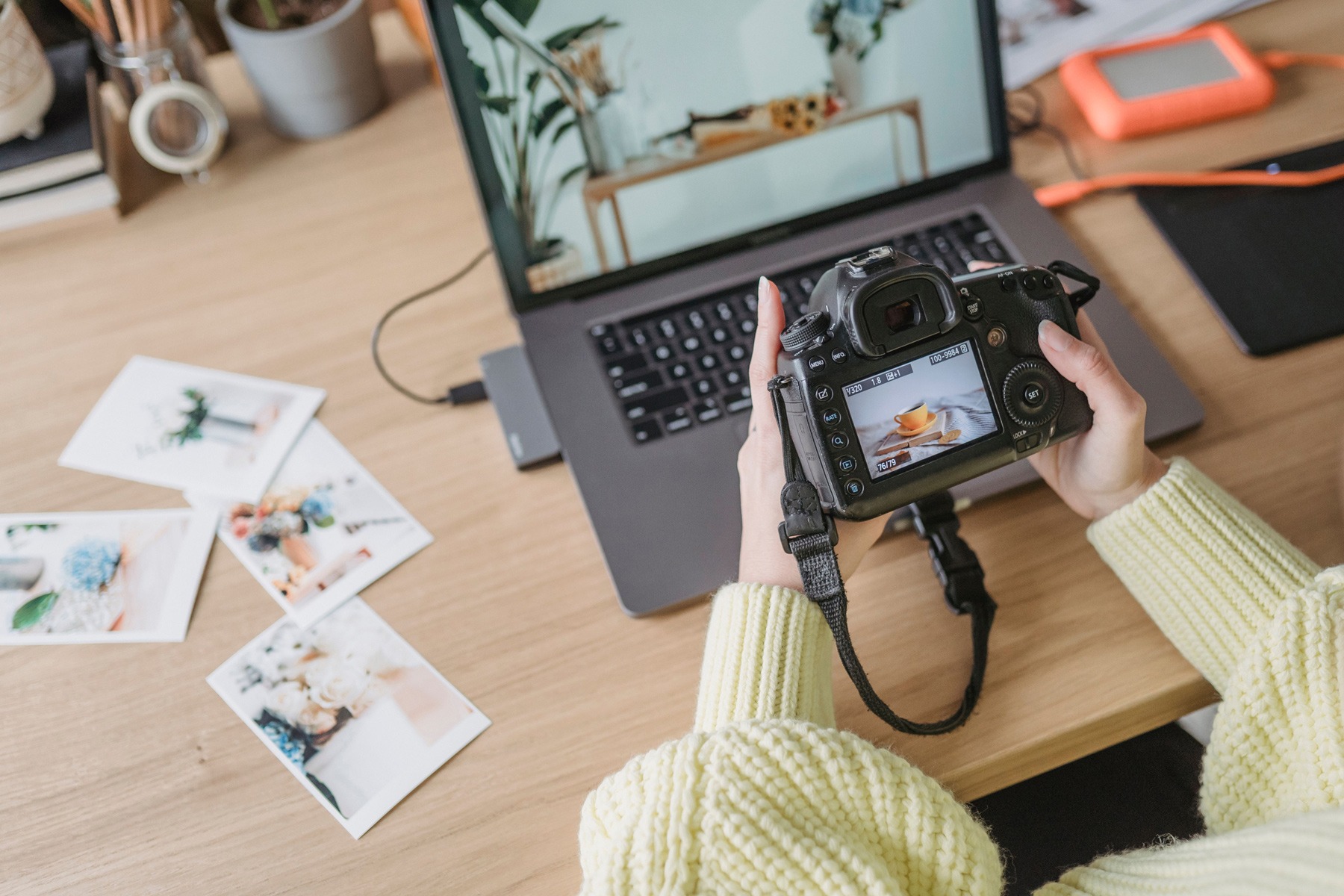 An overhead view shows a person in a yellow knit sweater holding a DSLR camera displaying a photo of a coffee cup on its screen, with a laptop and printed photos scattered on the wooden desk.