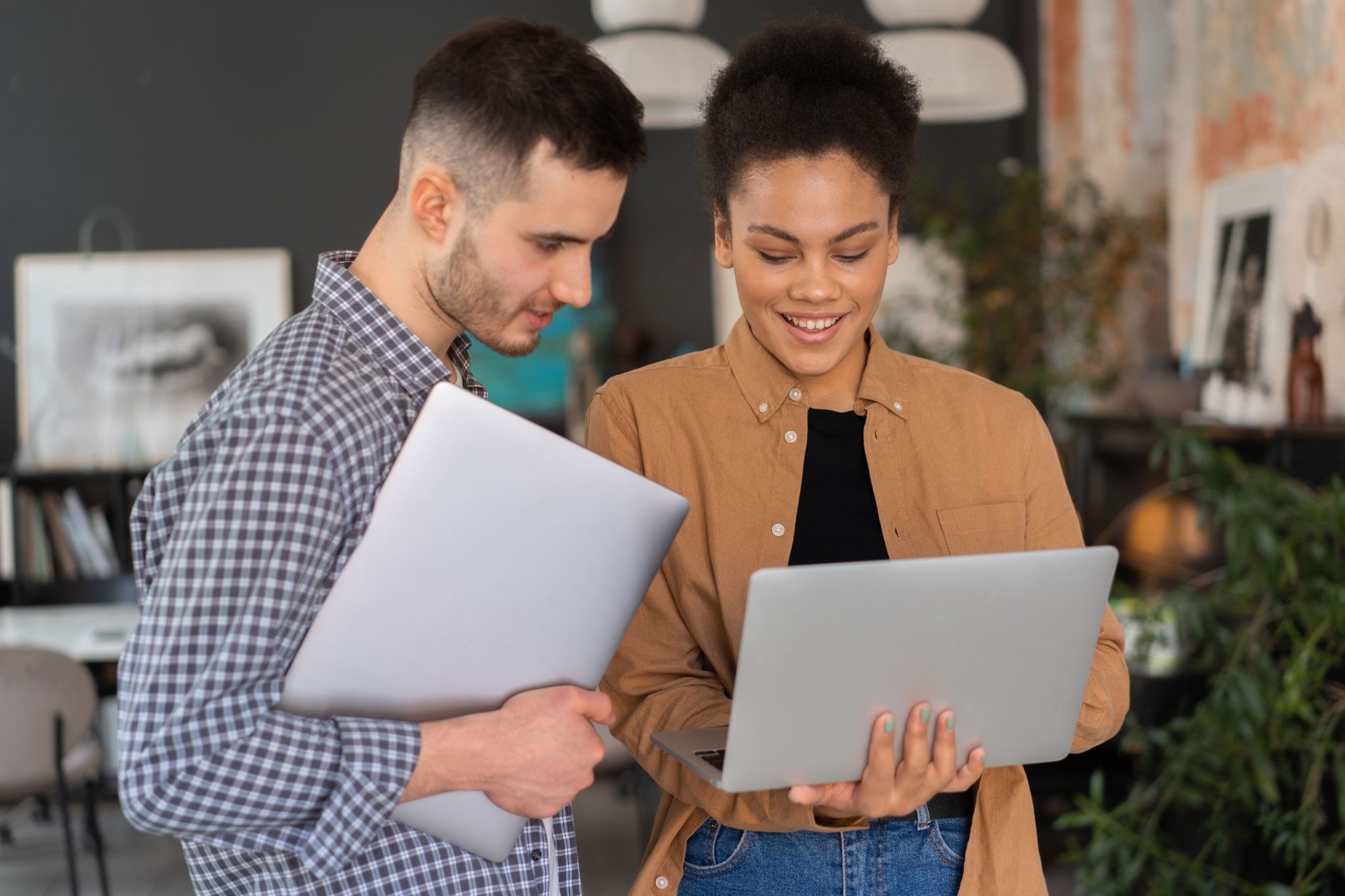 A smiling woman in a brown button-down shirt holds an open laptop while a man in a plaid shirt stands beside her holding a closed laptop and looking at her screen in a modern office setting.