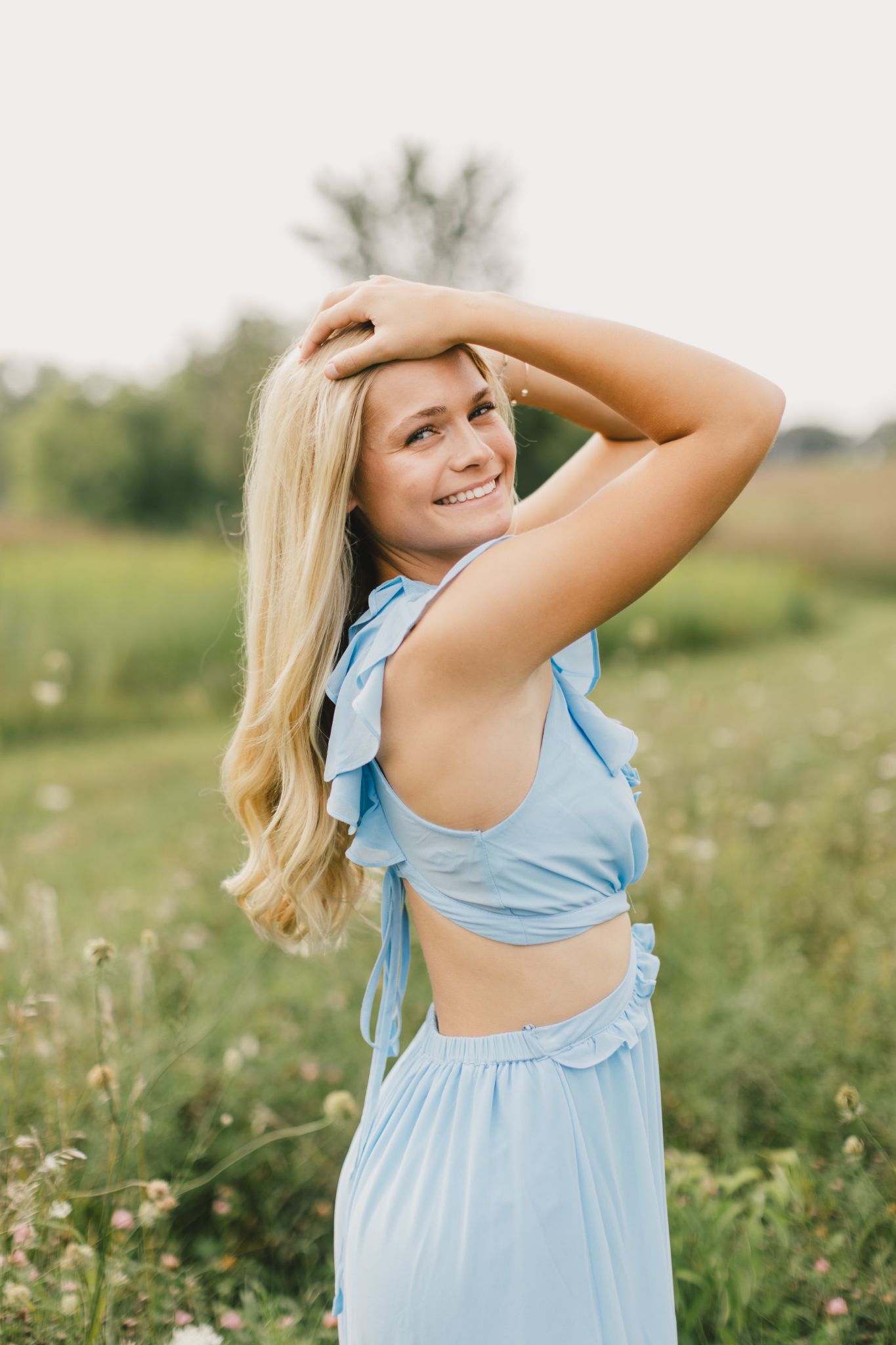 Smiling blonde woman posing outdoors in a blue outfit surrounded by wildflowers