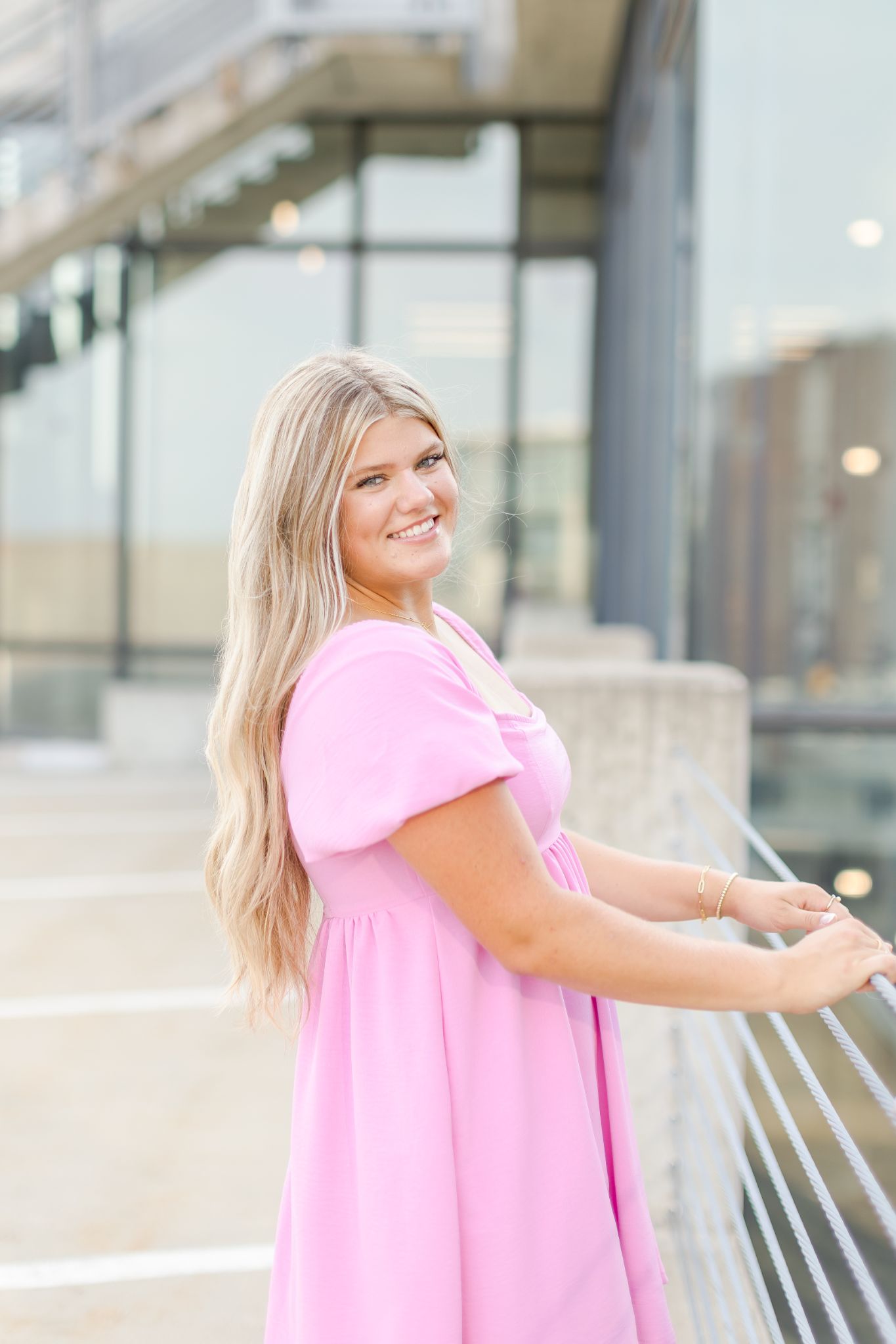 Smiling woman in pink dress leaning against a railing in a modern glass building