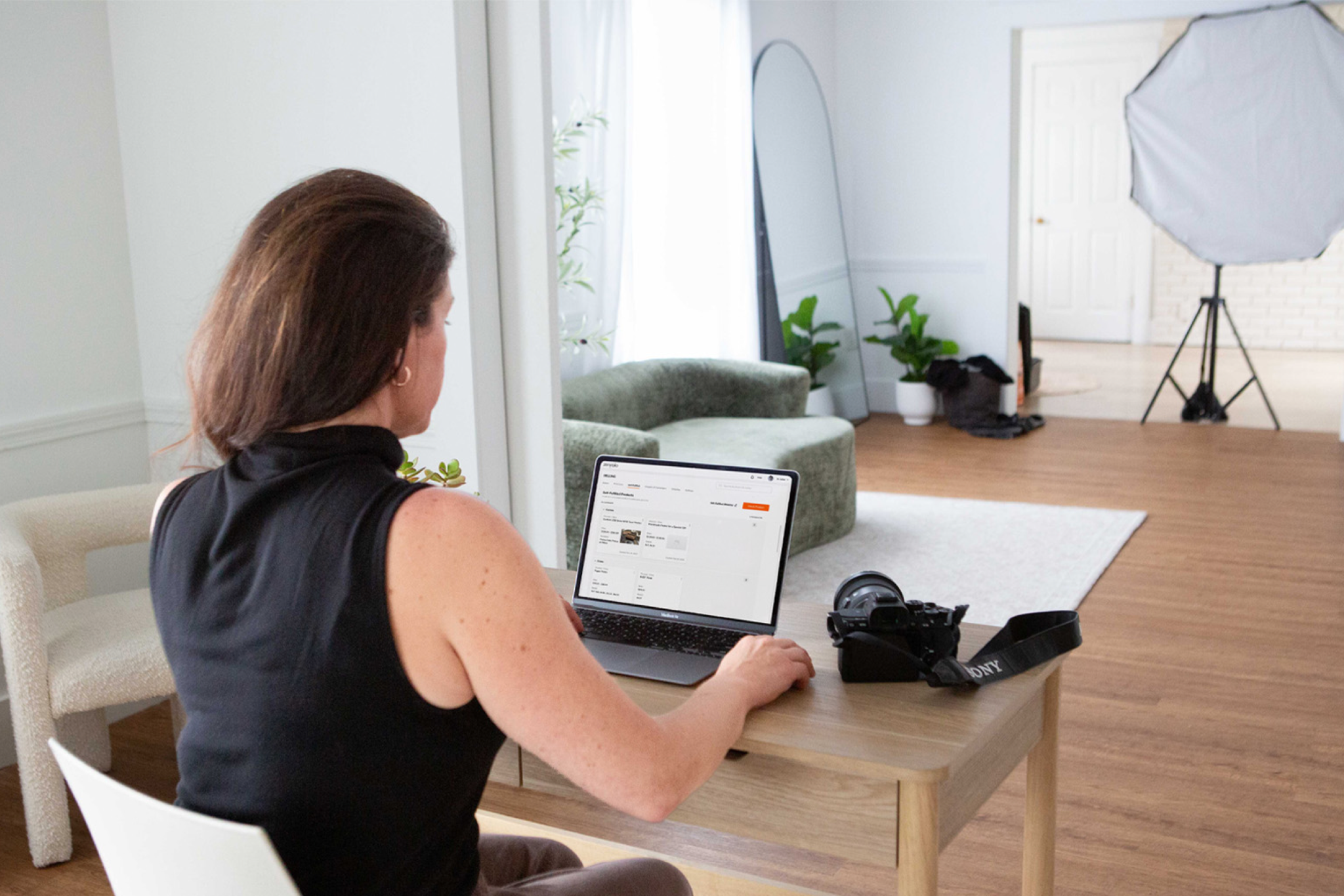A woman with brown hair sits at a wooden desk working on a laptop displaying a website dashboard, with a professional DSLR camera sitting next to her in a bright home photography studio setup.
